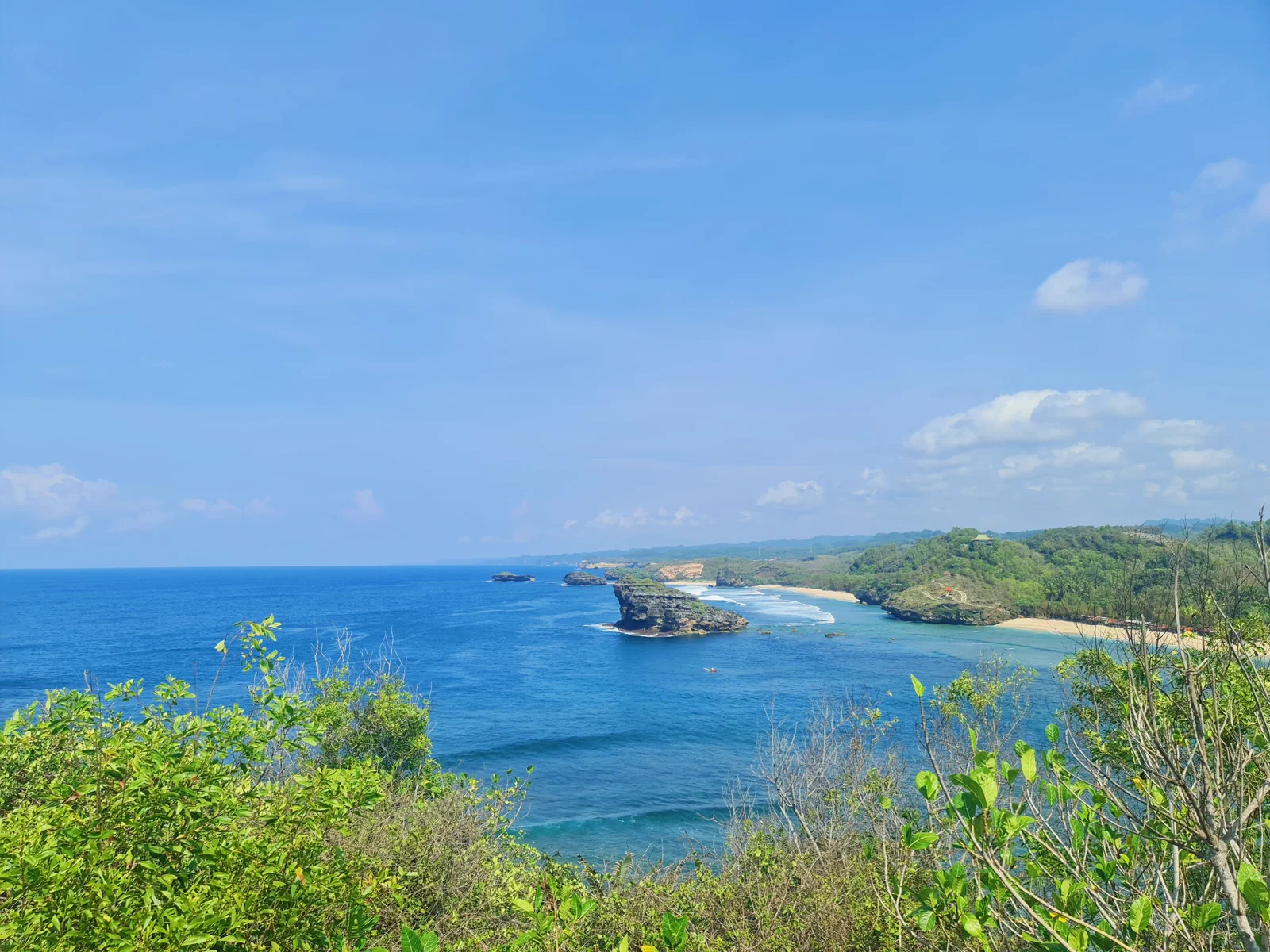 Teluk berair biru dengan tanjung karang berumput dan garis pantai berpasir putih dilihat dari bukit di atas Pantai Watu Karung