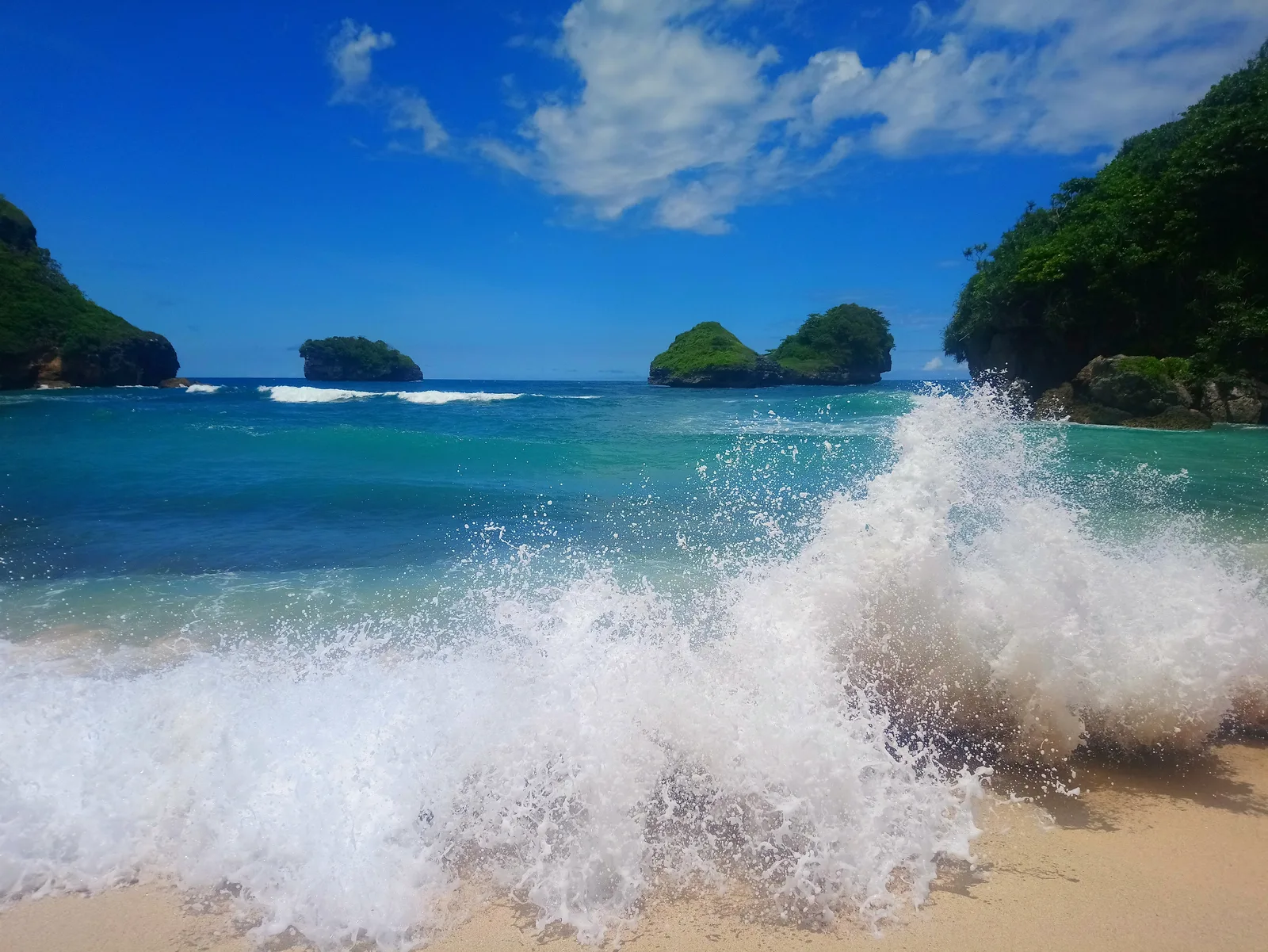 Ombak pecah di pasir Pantai Goa Cina dengan dua pulau karang bervegetasi dan tebing hijau di kedua sisi bingkai