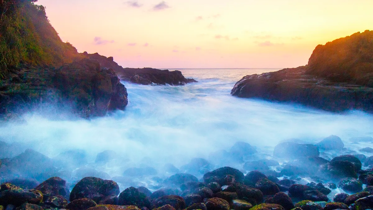 Matahari terbenam di Pantai Wediombo, Gunungkidul, dengan ombak berbuih halus di antara batu karang