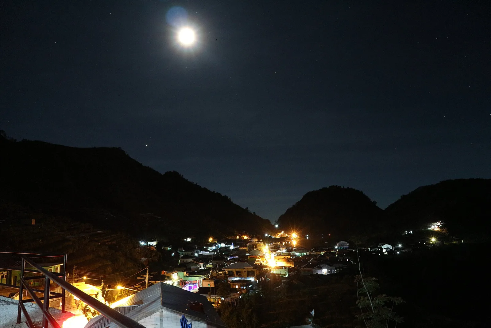 Permukiman di dataran tinggi Dieng menyala di bawah cahaya bulan, dilihat dari lereng bukit pada malam hari