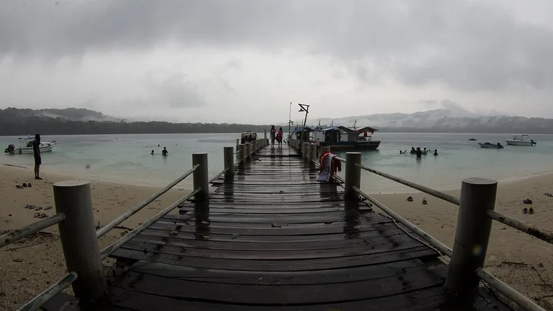Dermaga kayu panjang Pulau Peucang menjorok ke laut dangkal dengan beberapa perahu dan pengunjung di air saat langit mendung