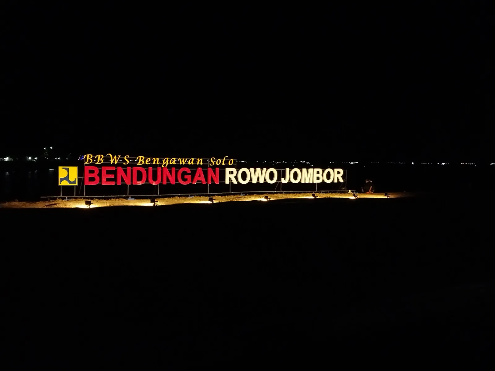 Papan nama bercahaya bertuliskan Bendungan Rowo Jombor BBWS Bengawan Solo pada malam hari di tepi waduk