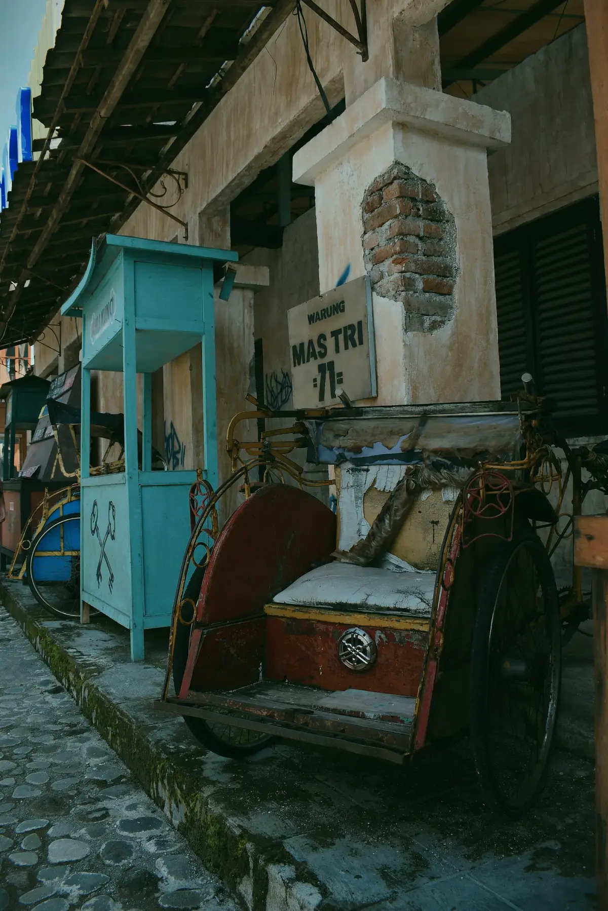 Becak tua dan gerobak dagangan kayu berdiri di lorong berbatu di antara bangunan set Studio Alam Gamplong