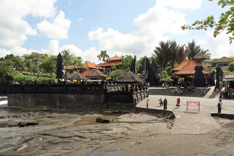 Pelataran kawasan wisata Pura Tanah Lot dengan bangunan beratap Bali dan pengunjung berjalan di tepi pantai berbatu
