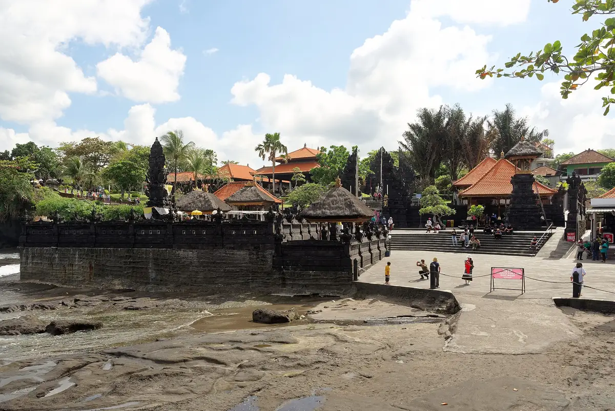 Pelataran kawasan wisata Pura Tanah Lot dengan bangunan beratap Bali dan pengunjung berjalan di tepi pantai berbatu
