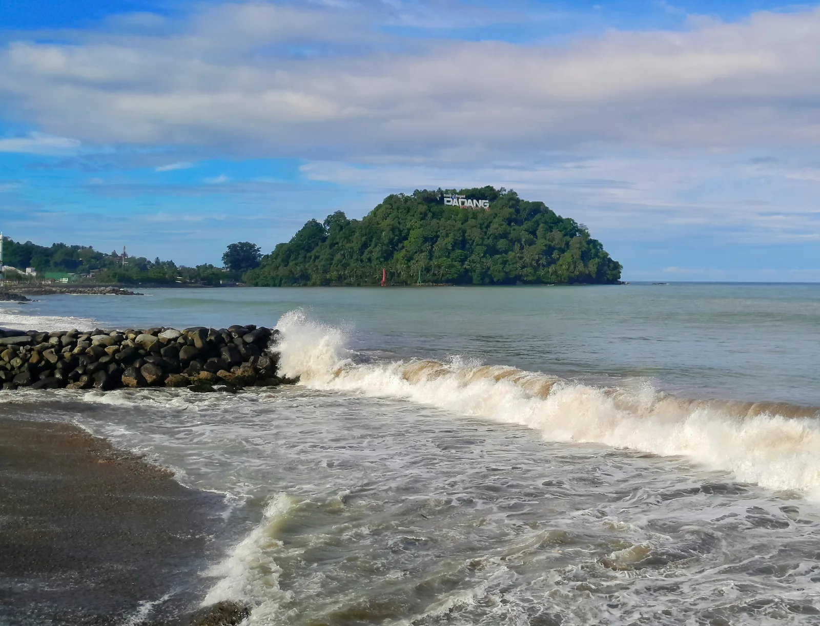 Ombak pecah di pemecah gelombang batu Pantai Padang dengan Gunung Padang yang berhutan di latar belakang