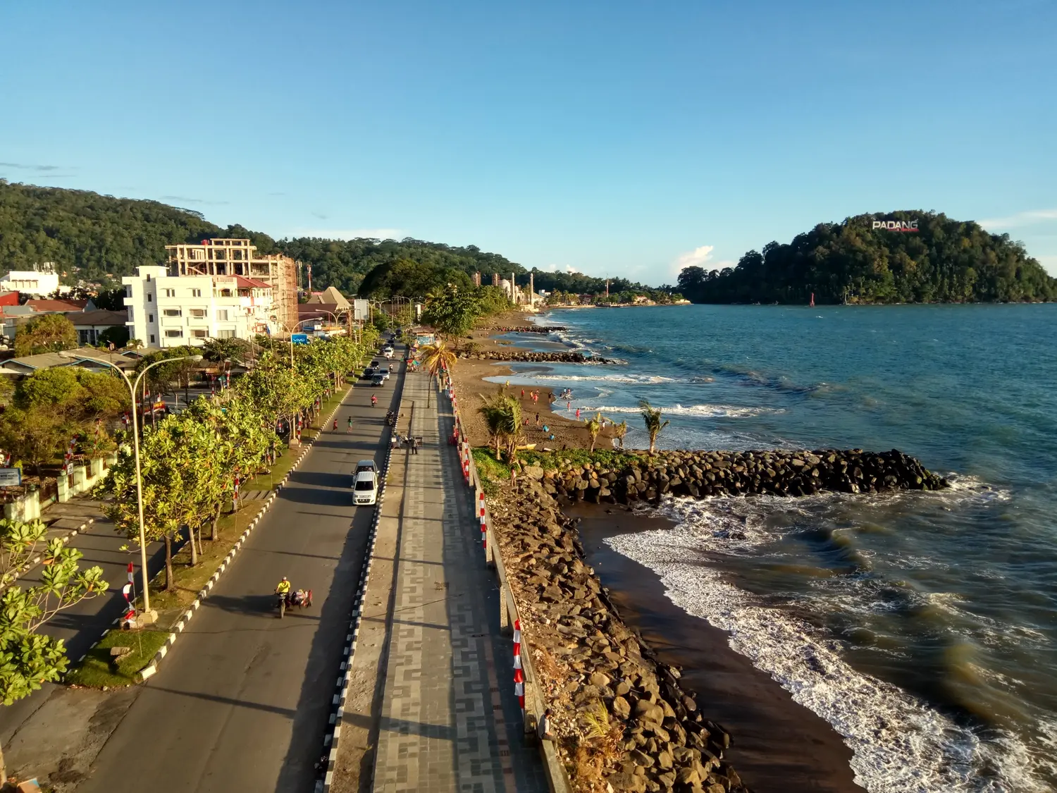 Jalan tepi laut dan trotoar lebar sepanjang Pantai Padang menjelang sore, dengan bukit bertuliskan PADANG di kejauhan