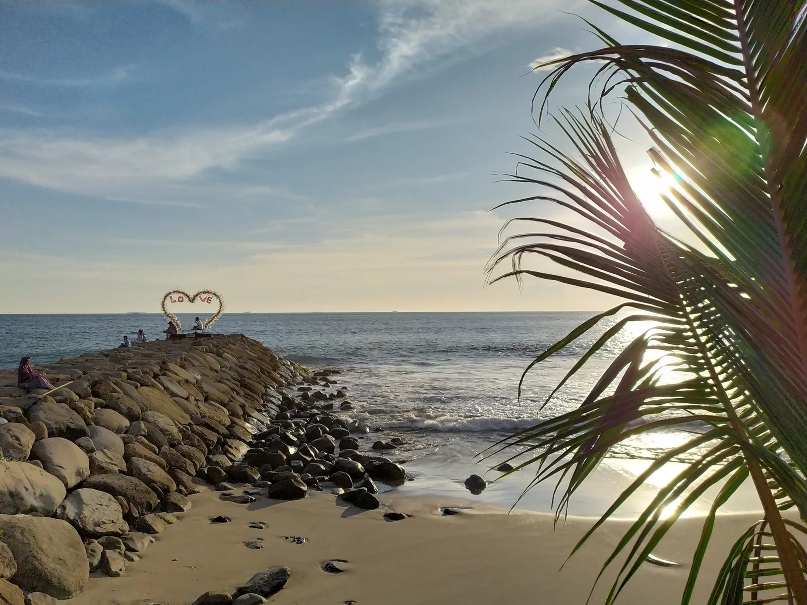 Ornamen berbentuk hati di ujung tanggul batu Pantai Padang saat senja dengan pelepah kelapa membingkai sisi kanan