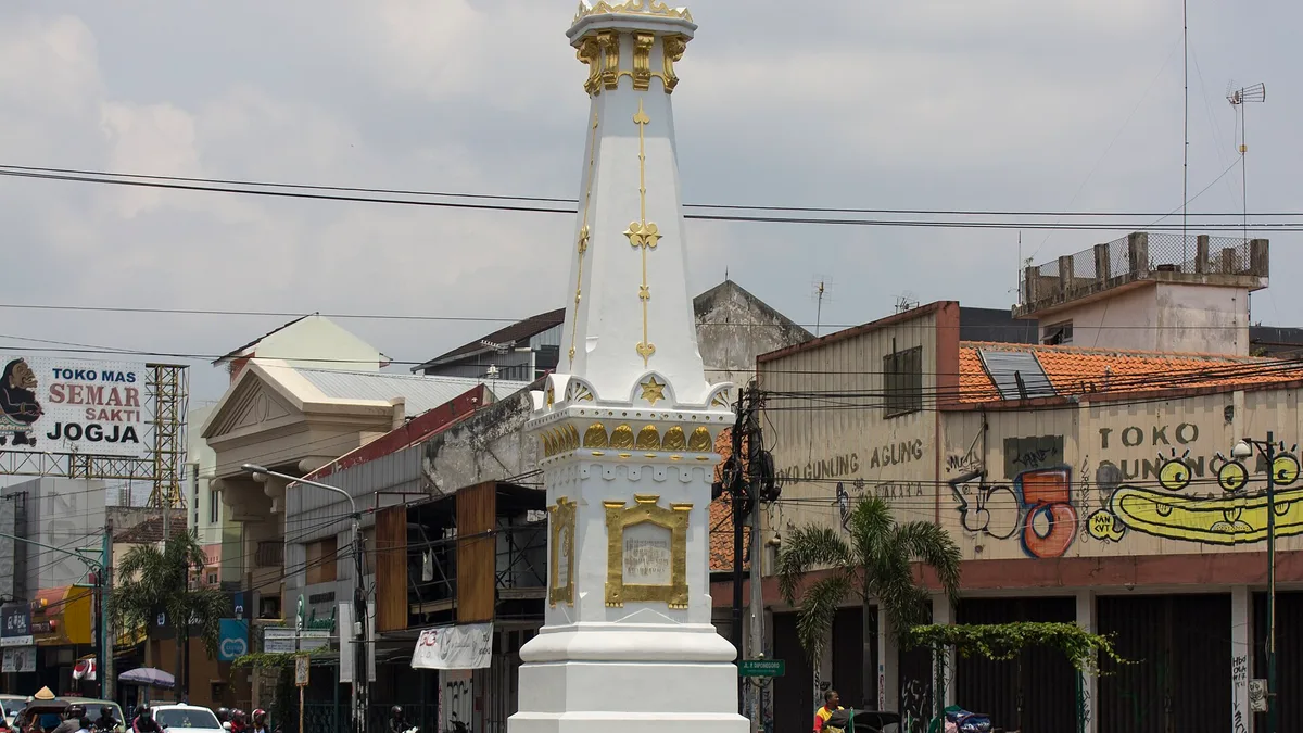 Tugu Yogyakarta berwarna putih emas berdiri di tengah persimpangan jalan dengan deretan pertokoan di belakangnya