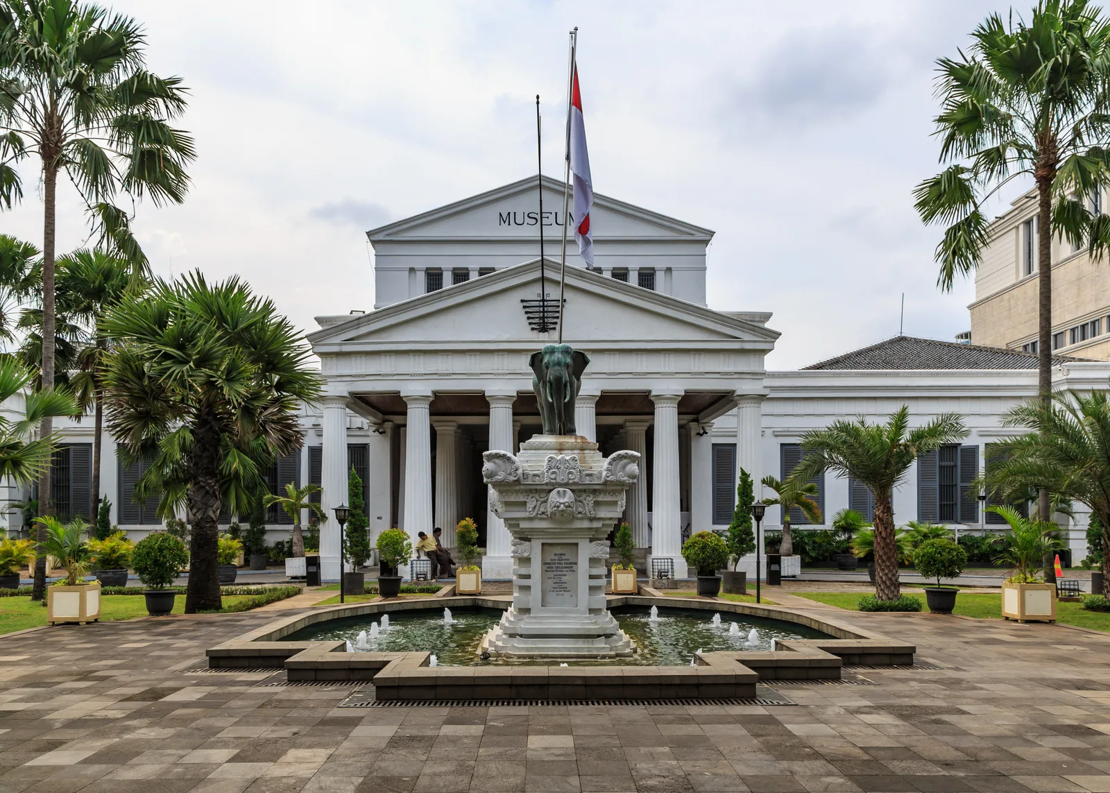 Fasad kolonial Museum Nasional Indonesia berkolom putih dengan patung gajah perunggu di atas air mancur di halaman depan