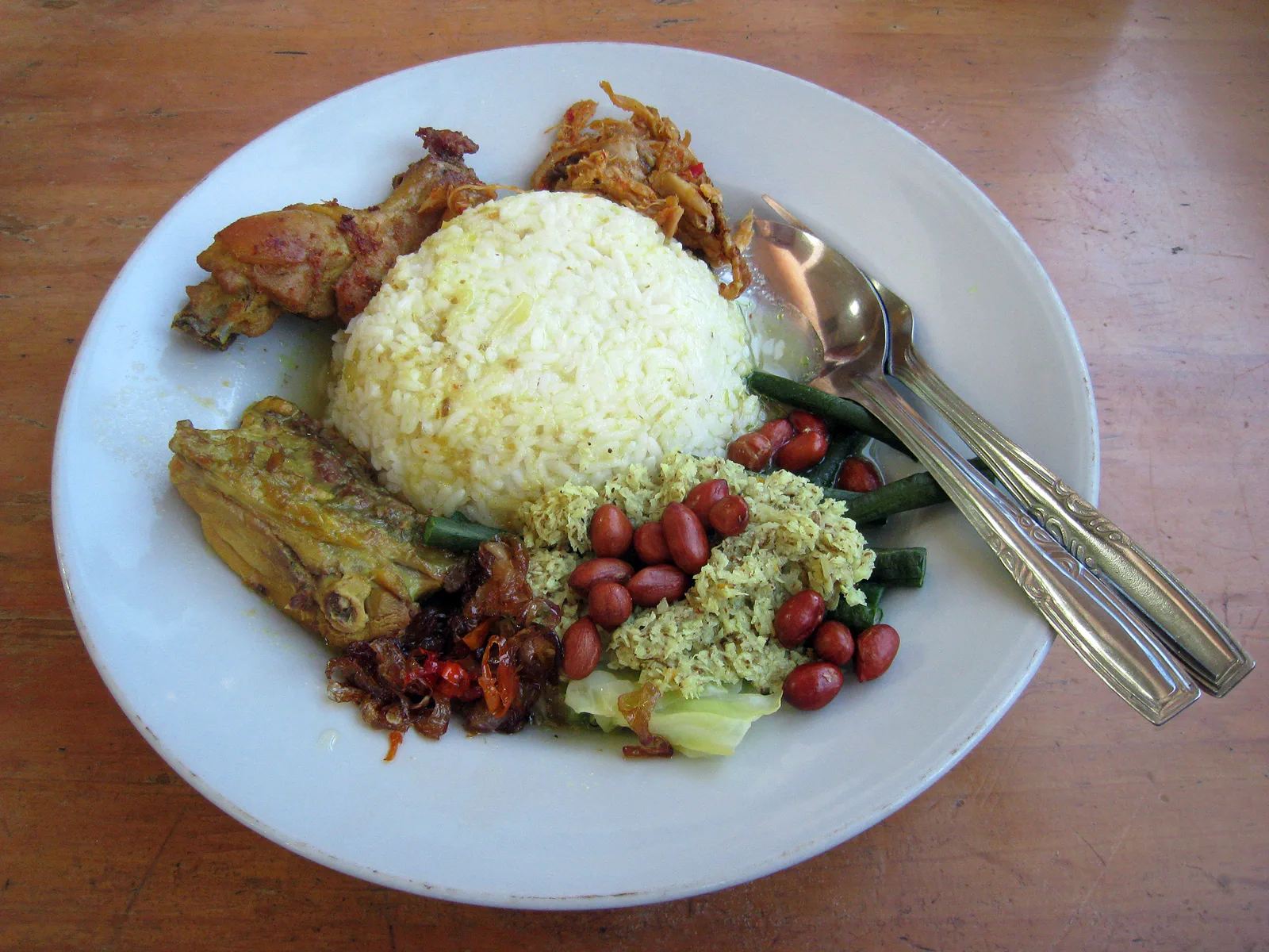 Sepiring nasi campur Bali berisi ayam betutu, urap kelapa parut, kacang tanah, buncis, dan sambal di atas meja kayu