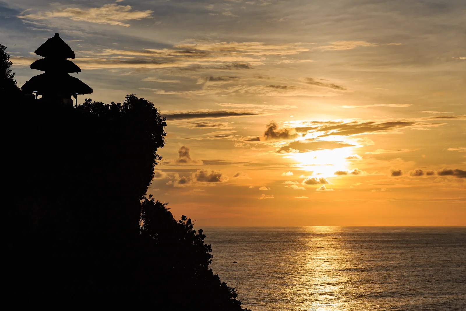 Siluet meru bertingkat Pura Luhur Uluwatu di ujung tebing dengan matahari terbenam di atas Samudra Hindia