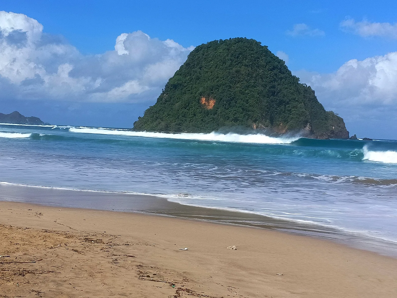 Bukit hijau berbentuk kerucut di lepas Pantai Pulau Merah, Banyuwangi, dengan ombak putih pecah di depannya dan hamparan pasir cokelat di latar depan