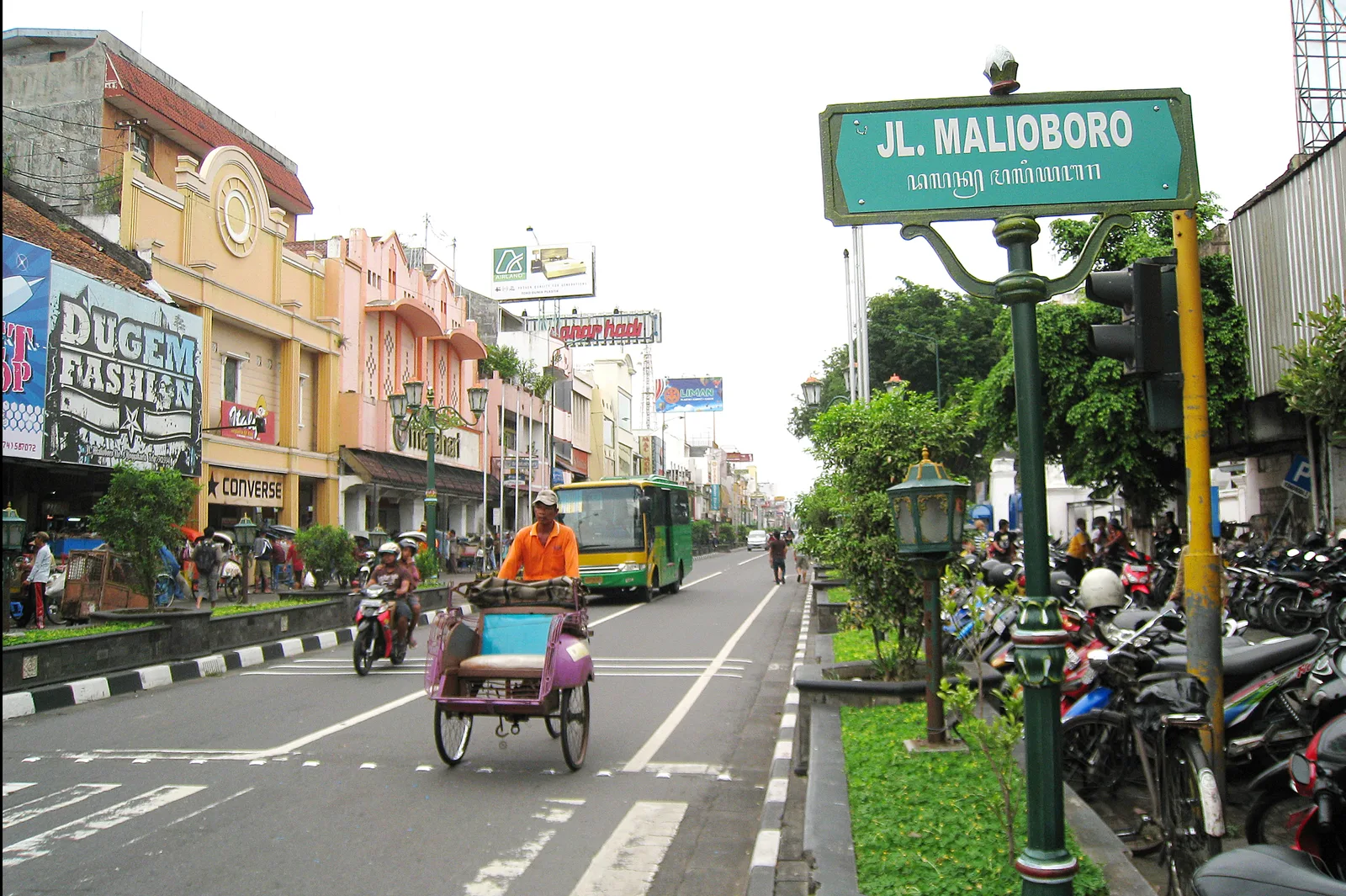 Papan nama Jalan Malioboro berbahasa Indonesia dan aksara Jawa, dengan becak, bus Trans Jogja, dan deretan sepeda motor terparkir