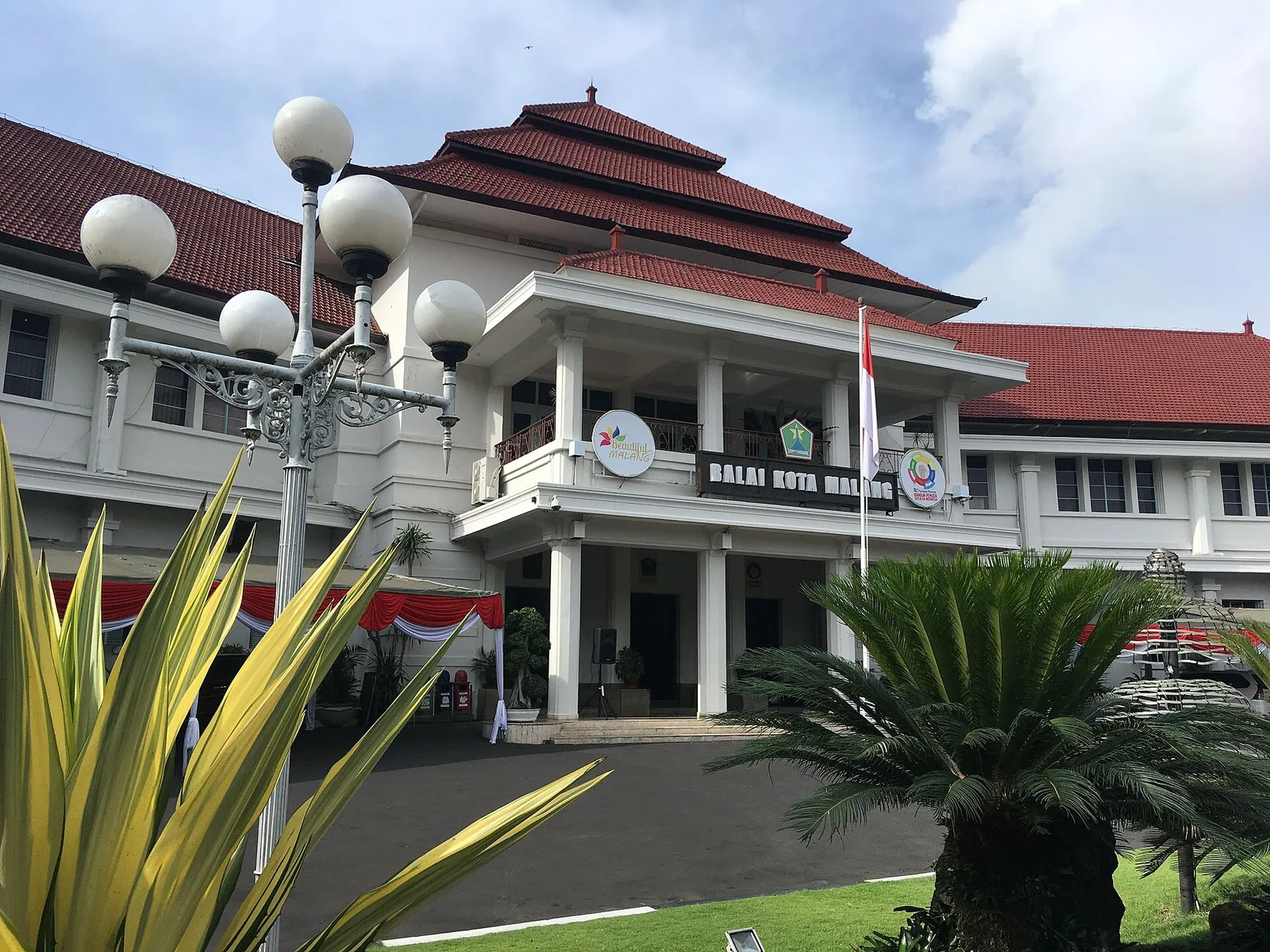 Gedung Balai Kota Malang bercat putih beratap joglo merah dengan lampu taman dan tiang bendera di halaman depan