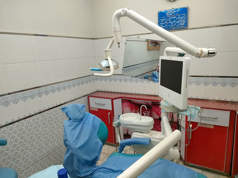 Kursi dokter gigi berlapis kain biru dengan lampu periksa, wastafel kumur, dan monitor di ruang praktik