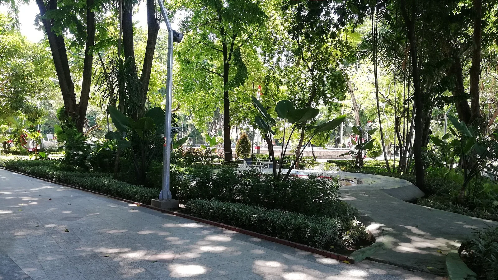 Jalan setapak berpaving di Taman Bungkul Surabaya dengan air mancur kecil dan pepohonan rindang di kanan kiri