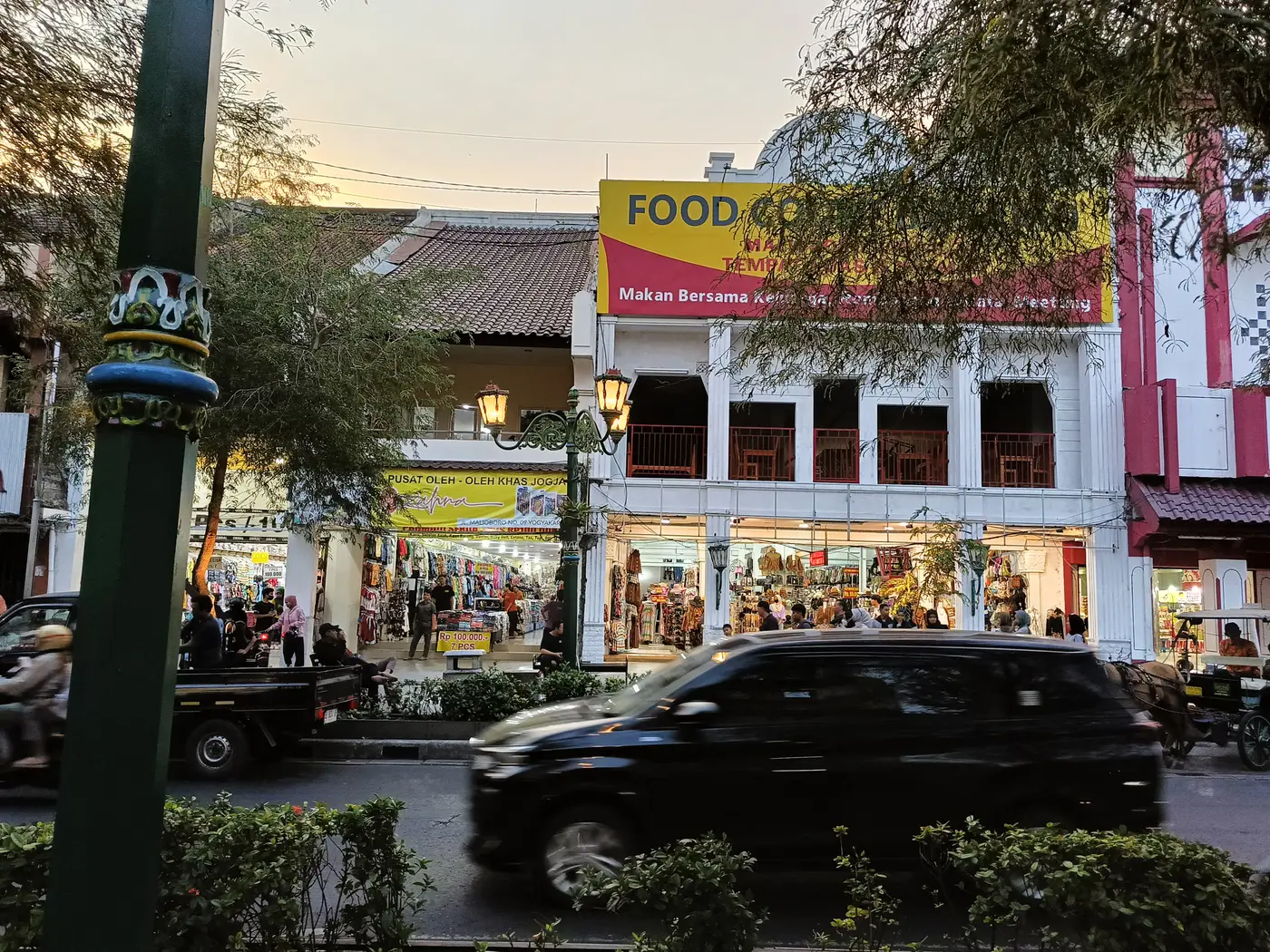 Deretan toko oleh-oleh dan bangunan lama di tepi Jalan Malioboro menjelang senja dengan tiang lampu jalan bergaya klasik