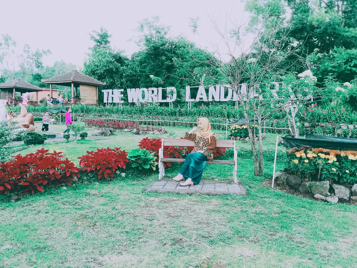 Tulisan THE WORLD LANDMARKS berdiri di antara petak bunga di taman Merapi Park, Jalan Kaliurang, dengan pengunjung duduk di bangku kayu