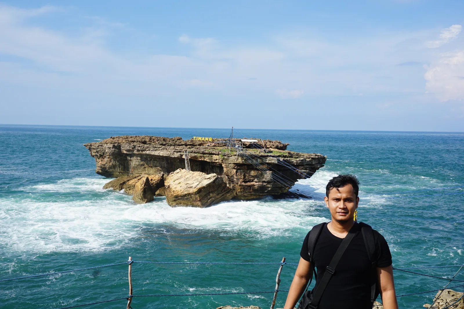Pulau karang bertuliskan TIMANG dikelilingi ombak putih, dengan tali gondola membentang dan seorang pengunjung berdiri di tepi tebing